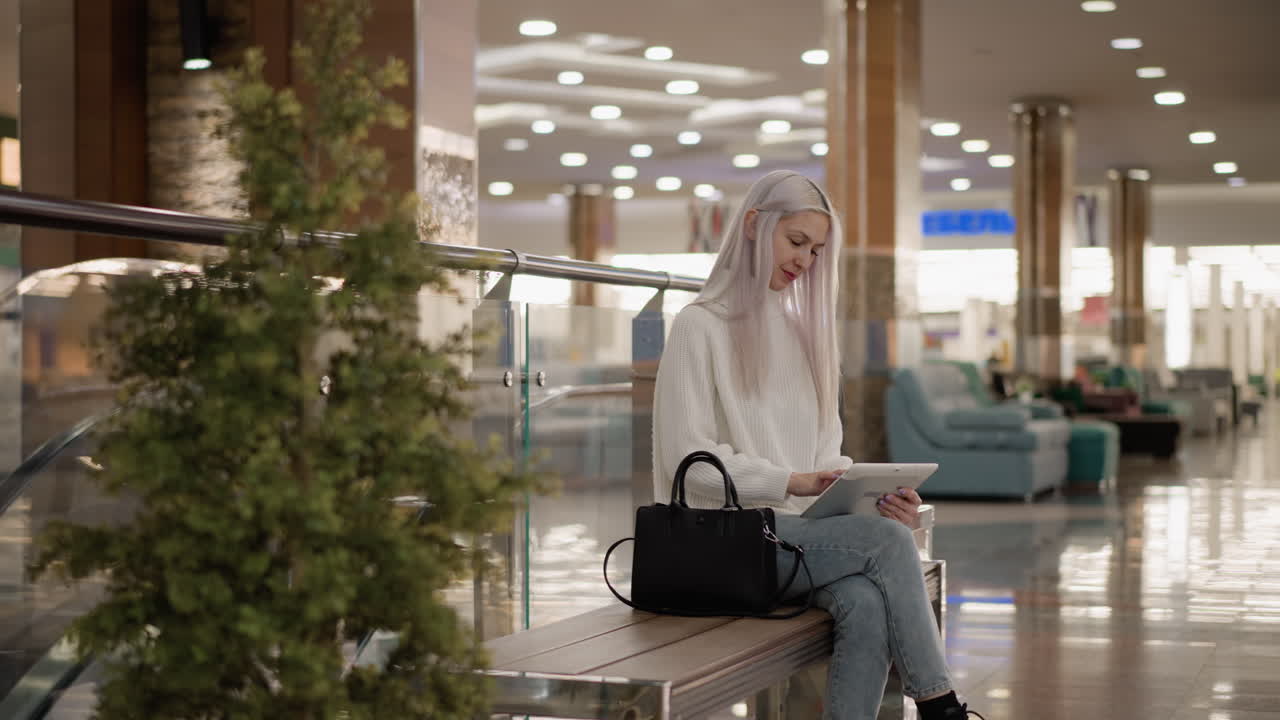 mindful woman seated on bench in bright mall hall tapping tablet screen with smile and eyes fixed working browsing content in relaxed modern interior lit by decorative lights glass railing escalator