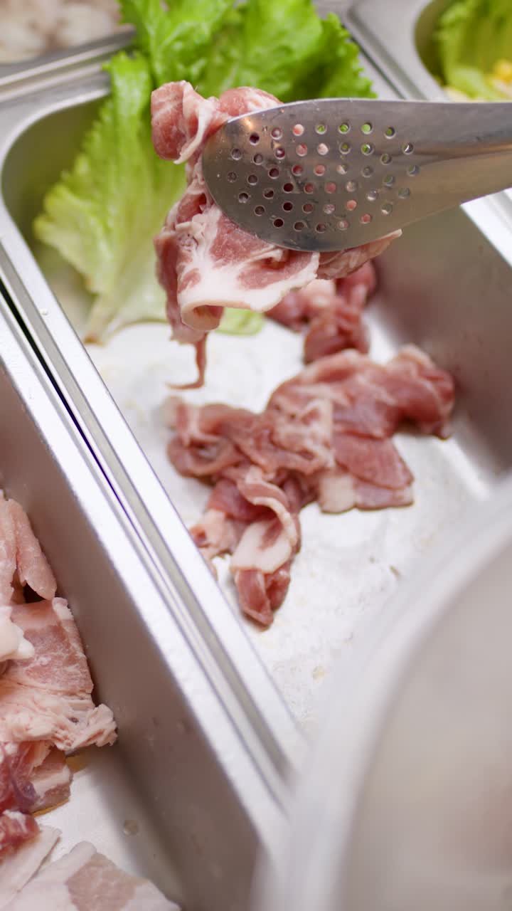 Hand uses tongs to pick raw sliced meat from buffet tray under bright, even lighting