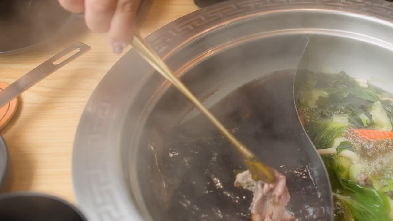 Hand dips sliced beef into steaming dual broth hotpot, surrounded by fresh vegetables, overhead view