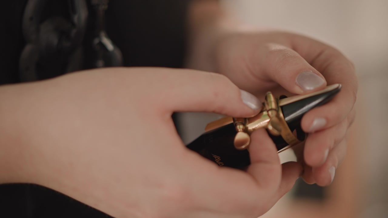 Primo piano di mani che montano la legatura e il bocchino di un clarinetto in uno studio accogliente. Un insegnante di musica prepara l'ancia con dita ferme, con una manicure curata visibile, eseguendo una regolazione mirata e lenta per un tono ottimale.
