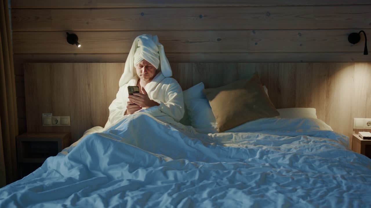 En person i badekåbe slapper af i sengen på en rustik hytte, fordybet i sin smartphone blandt varm belysning og behageligt sengetøj. Scenen udstråler afslapning og komfort.