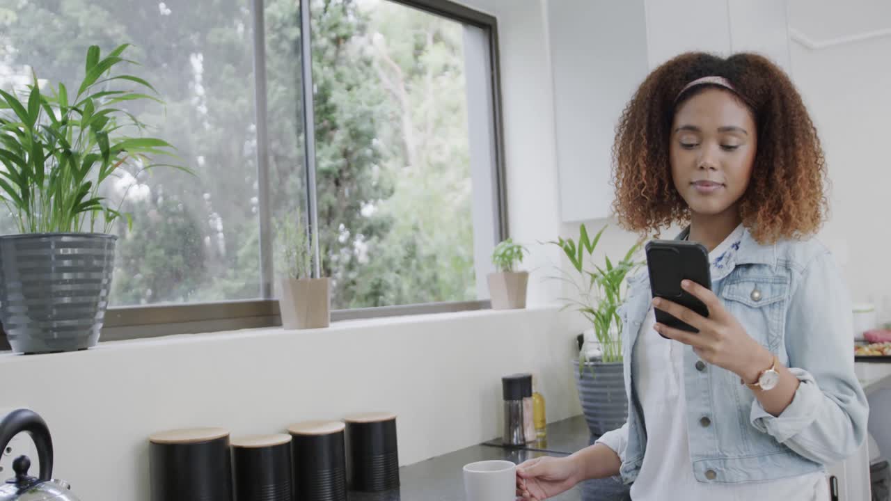 Happy biracial woman talking on smartphone and drinking coffee in kitchen, slow motion