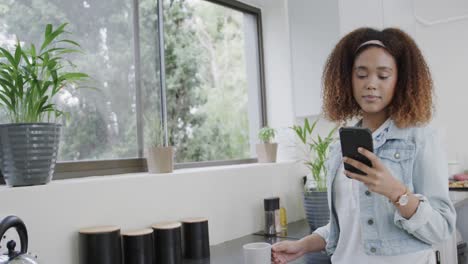 mujer biracial feliz hablando en el teléfono inteligente y bebiendo café en la cocina, cámara lenta