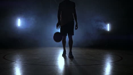 Jugador-De-Baloncesto-Masculino-Moviéndose-En-El-Humo-Ante-La-Cámara-Golpeando-La-Pelota-Sobre-El-Suelo-De-Parquet-En-Cámara-Lenta-Con-Steadicam