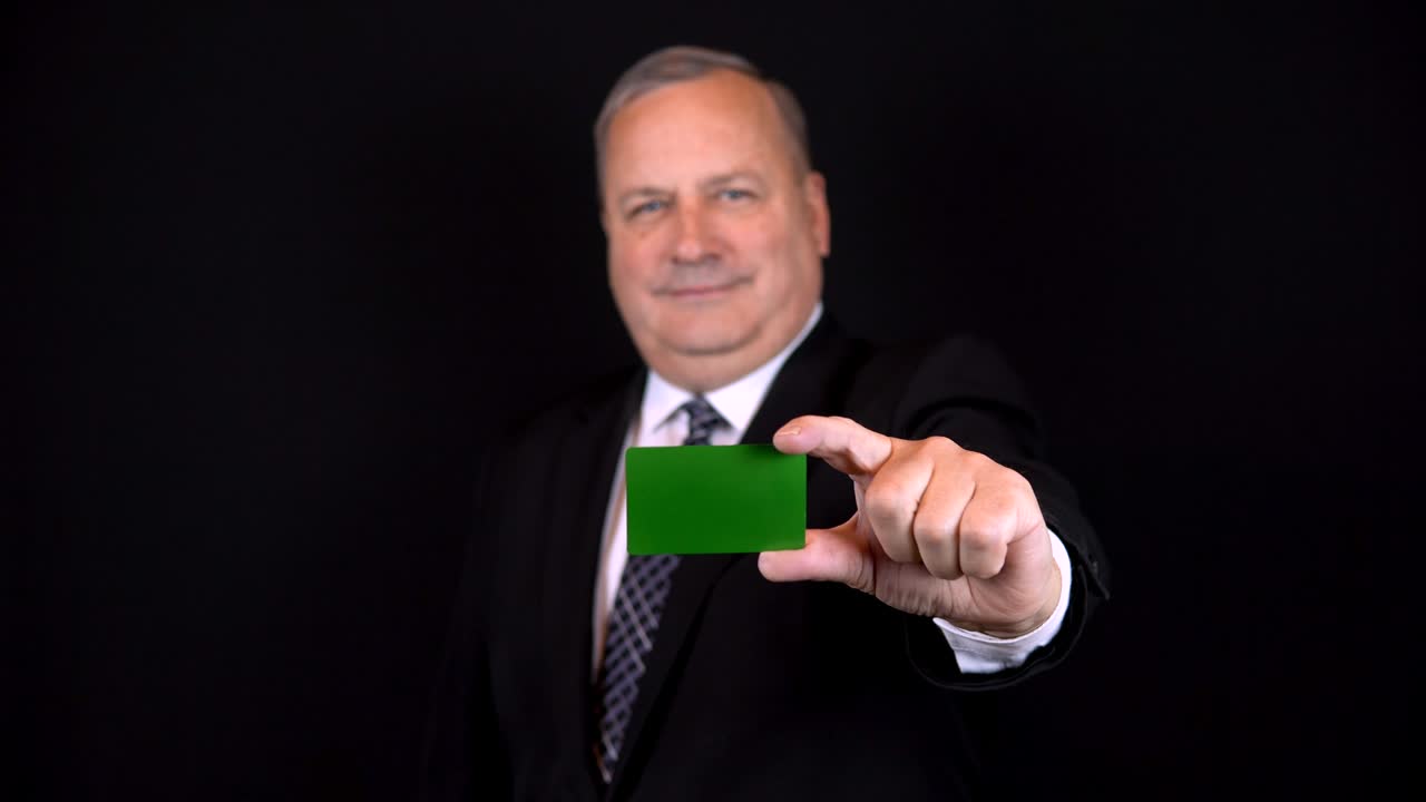Old businessman in a suit holds a bank card in his hand. Chromakey green card. Isolated black background.