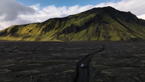 aerial push in shot of a car parking on a black volcanic road with a mountain in the background, on a sunny day with some clouds