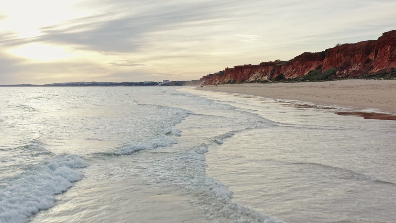 Low Aerial gliding shot over ocean waves along Sandstone cliffs and sandy beach at Algarve Coastline