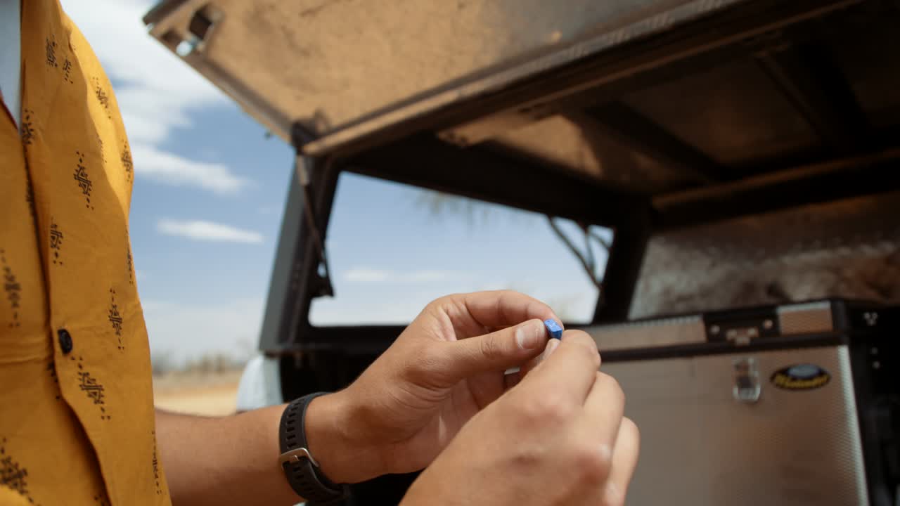 Dynamic tracking close-up shot of a Caucasian male tourist in Africa as he inspects and inserts the fuse of a camping fridge in the back of a off-roading vehicle.