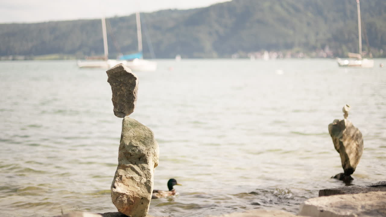 A peaceful scene showing a balanced stone sculpture near a calm lake as ducks gently swim across the water.