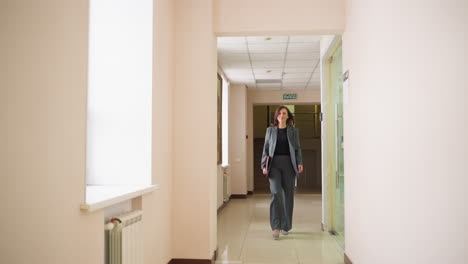 Confident businesswoman walking purposefully down modern hallway, holding red folder, dressed in professional attire, showcasing determination and focus, ideal for corporate