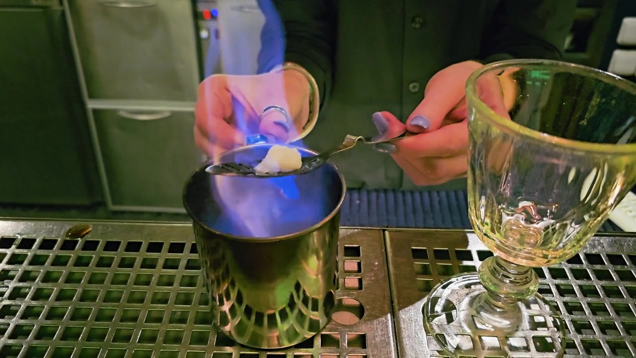 Primo piano di un barista che prepara l'assenzio in un cocktail bar, con fuoco blu, zolletta di zucchero, cucchiaio forato, tazza di metallo, calice di vetro, bancone in acciaio inossidabile e atmosfera da vita notturna.