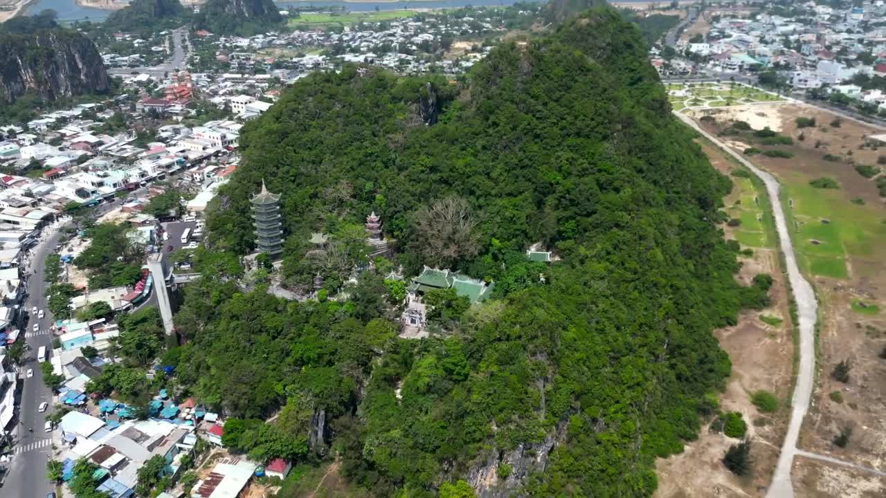 Nestled in greenery, an ancient temple hides beneath a forested hill, surrounded by a modern Vietnamese cityscape where spirituality, history, and urban life meet.
