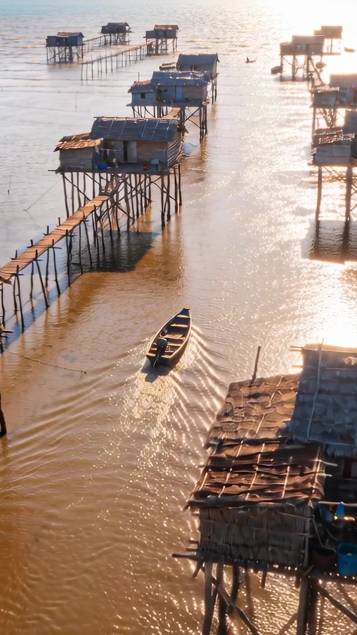 Video verticale: Drone in panoramica che mostra una barca che si muove lungo un estuario verso case su palafitte, con la scia
