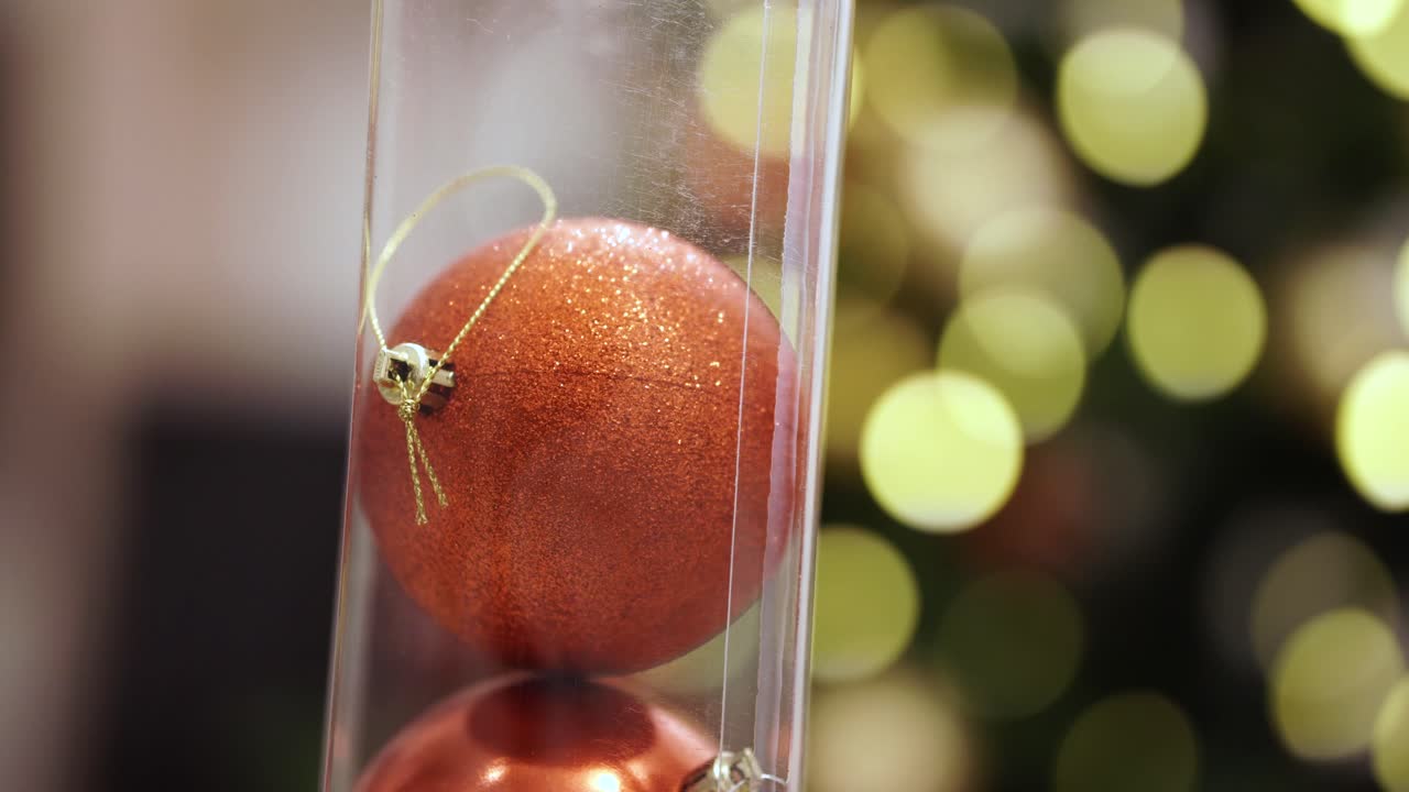 반짝이는 빨간색 크리스마스 공이 보케 배경에 노란색 조명이 있는 투명한 플라스틱 용기에 떨어집니다.