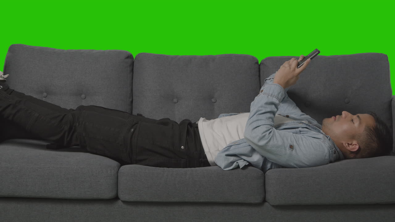 녹색 스크린을 배경으로 휴대폰을 보며 소파에 누워 있는 청년