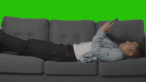 Young Man Lying On Sofa Looking At Mobile Phone Against Green Screen