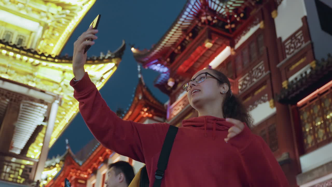Mulher asiática tira selfie em templo iluminado por lanternas à noite, jovem turista asiática com moletom vermelho segurando um smartphone, enquadrando o retrato contra telhados de pagode ornamentados e luzes douradas, estudante de arquitetura e rua