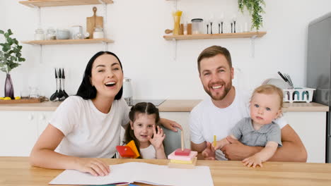 happy young parents sitting at table in kitchen playing with their two children while looking at camera during a video call