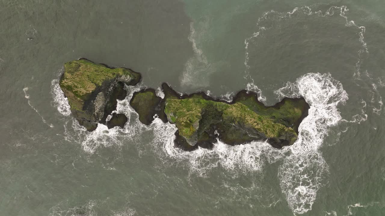 Aerial shot of a moss-covered rocky islet surrounded by crashing waves near Reynisfjara beach in southern Iceland, showcasing the rugged coastal landscape.