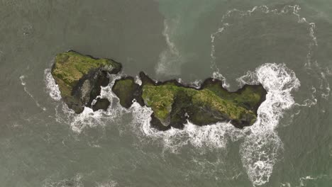 aerial shot of a moss-covered rocky islet surrounded by crashing waves near reynisfjara beach in southern iceland, showcasing the rugged coastal landscape.