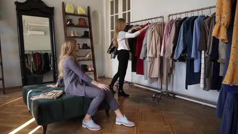 Long-Haired-Young-Blonde-Female-Is-Trying-A-New-Red-Coat-While-Standing-Near-The-Racks-With-Variety-Of-Outfits-On-Hangers