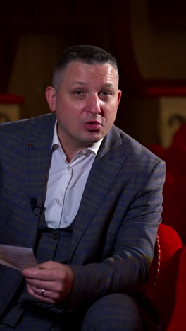 Man actor gives interview while sitting on red velvet chair in classic theater. Empty theter before evening performance. Vertical video