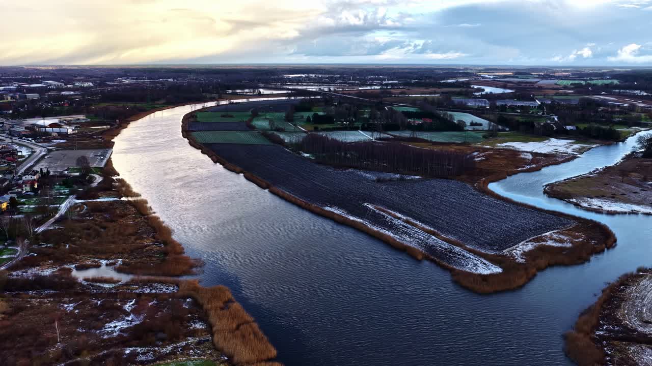 En snoet flod løber gennem marker og vådområder under en dramatisk himmel – droneoptagelse