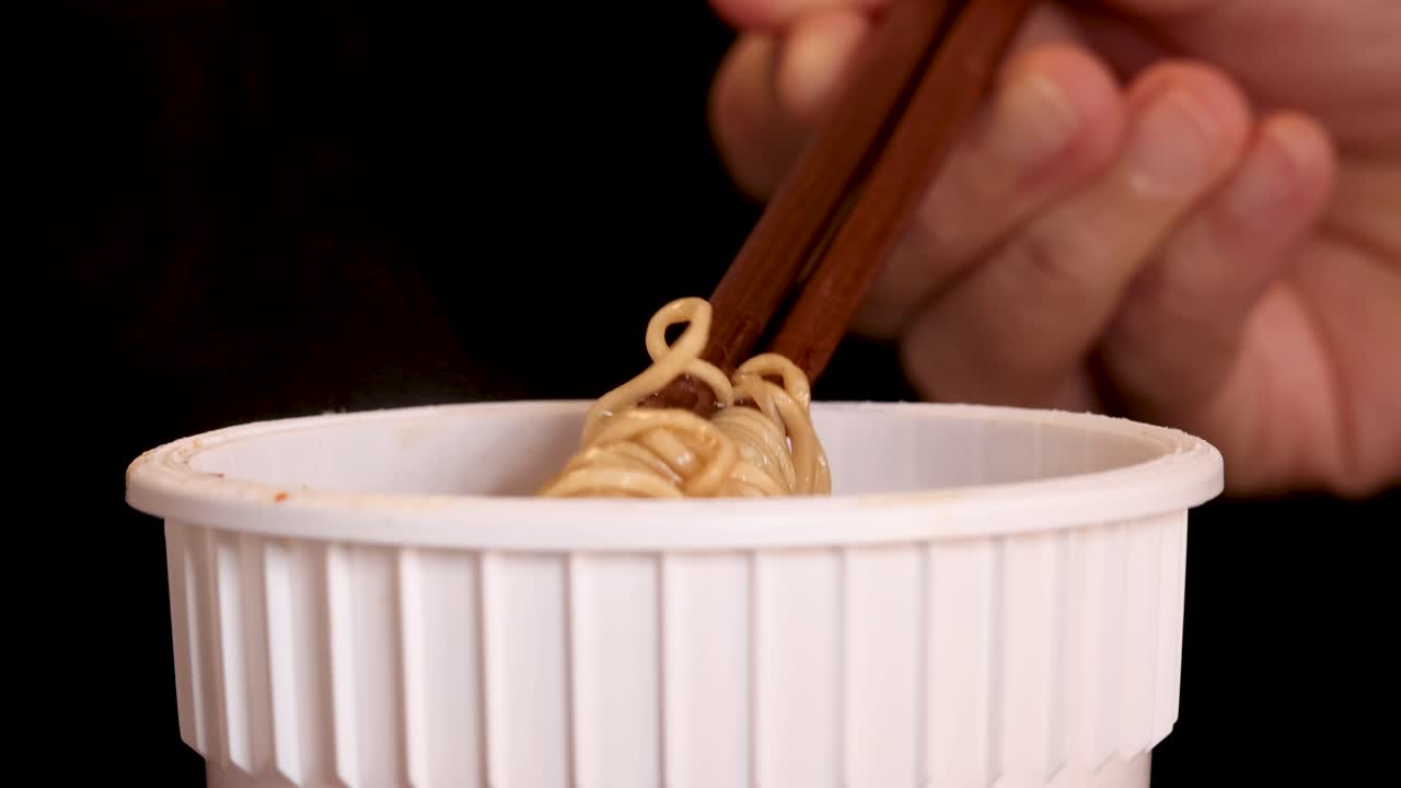 A hand skillfully uses chopsticks to lift cooked instant noodles from a white cup against a dark background, with warm, even lighting and close-up framing