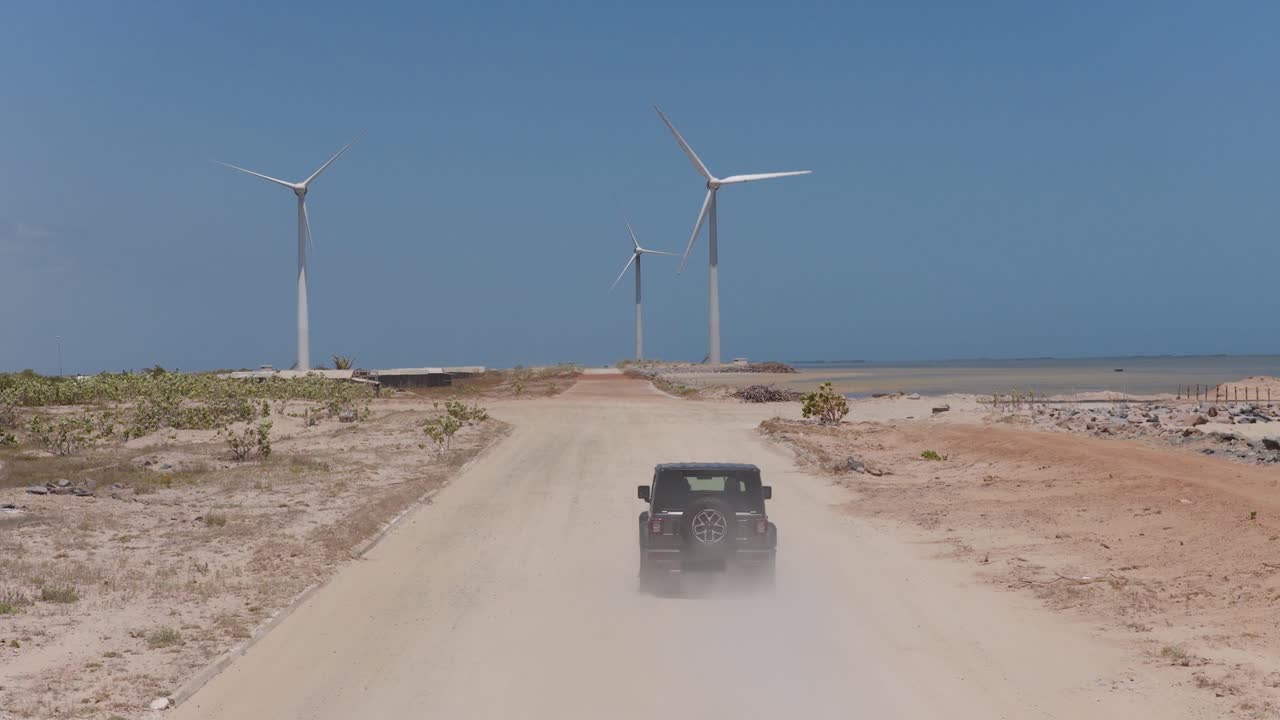 Eine professionelle Kamerafahrt folgt einem dunklen Geländefahrzeug, das eine sandige Küstenstraße entlangfährt. Moderne Windkraftanlagen ragen vor einem strahlend blauen brasilianischen Himmel über den Horizont.
