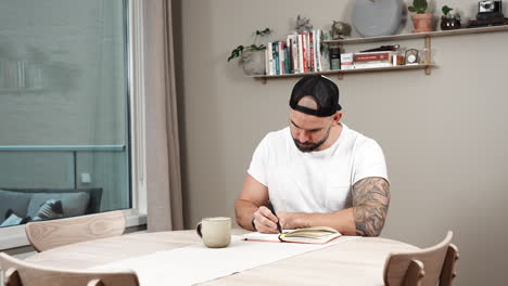 bearded and tattooed man writing diary on the table at home