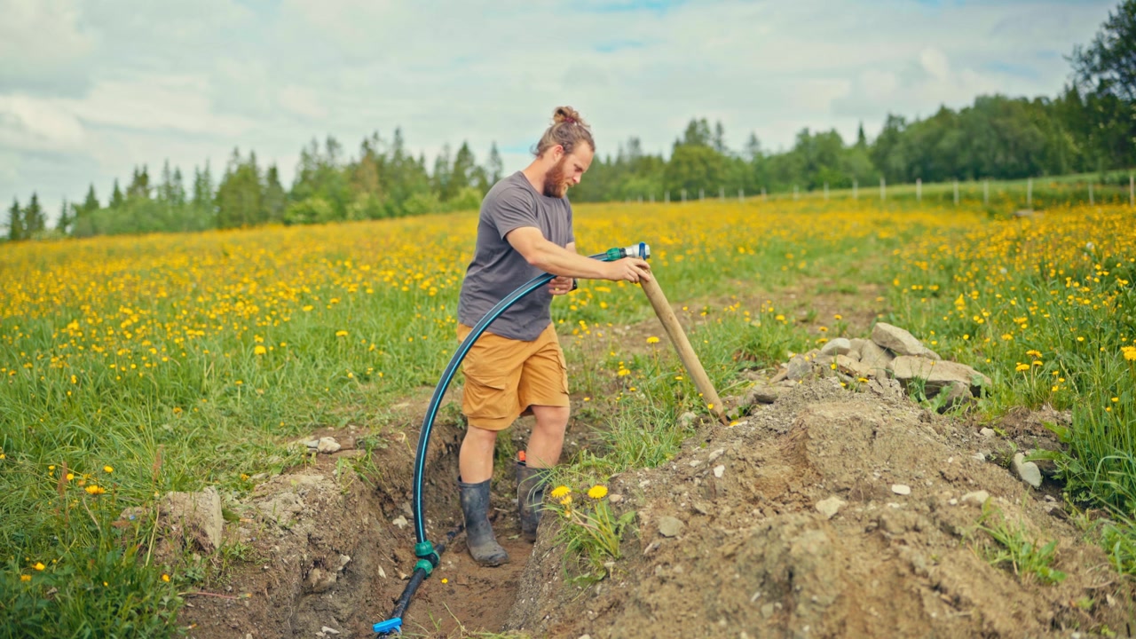 Osoba instalująca podziemny system wodny na farmie w Norwegii, ukazująca montaż rur i pracę ręczną w wiejskim środowisku rolniczym.