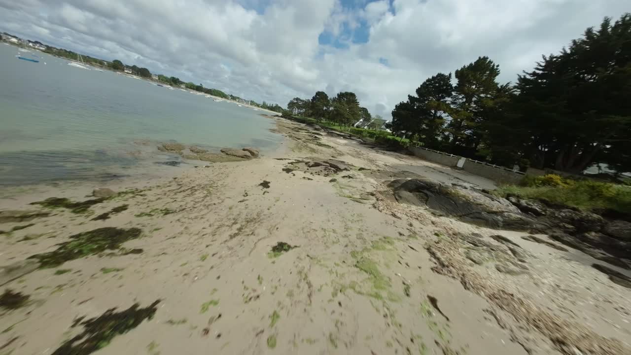 FPV drone twists and descends close to shoreline, flying toward sea water above sandy beach in Concarneau, Brittany, France, beauty of coastal landscape.