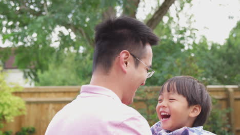 asian father lifting laughing son in  air as they play game in garden together