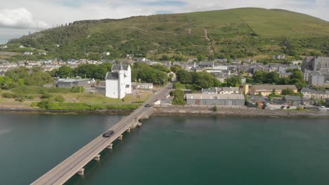 aerial camera follows automobiles traveling over a narrow bridge,past an unusual castle structure into a small seashore town