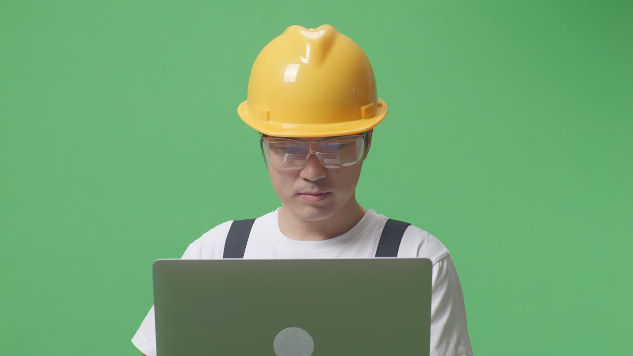 Close Up Of Asian Man Worker Wearing Goggles And Safety Helmet Using A Laptop While Standing In The Green Screen Background Studio