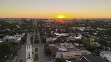 Dolly-Volando-Sobre-La-Iglesia-Neogótica-Y-La-Calle-Principal-En-La-Ciudad-Rural-De-Santa-Elisa-Al-Atardecer-Con-La-Puesta-De-Sol-En-El-Horizonte,-Entre-Rios,-Argentina