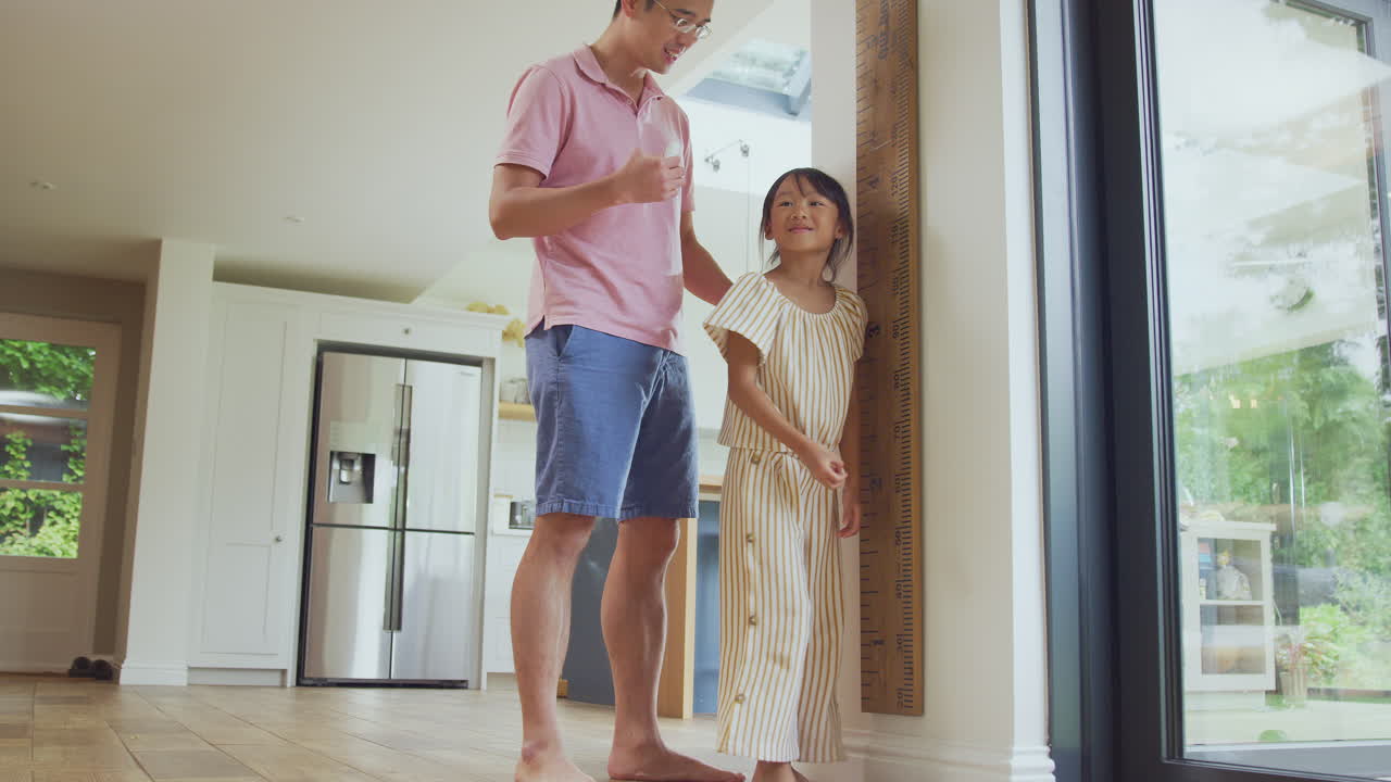 Asian Father Measuring Daughter On Wall Scale At Home As She Stands On Tip Toes