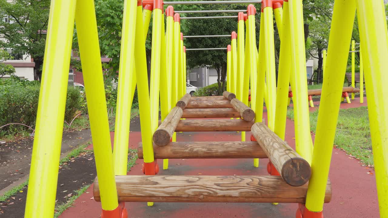 A low-angle shot of a wooden and metal jungle gym bridge in a park. The red safety matting is visible