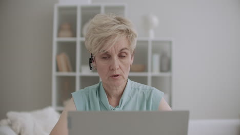 aged blonde woman is consulting online using video call and headphones talking and looking at screen of laptop