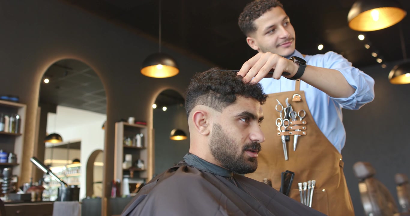 Barber kæmmer og trimmer siddende kunde, der ønsker en klipning, i salonen med kam, klippemaskine og forklæde