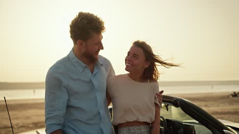 portrait: a bearded man in a blue shirt and a blonde girl in a white top are hugging and looking at the camera on the river bank during a bright sunset