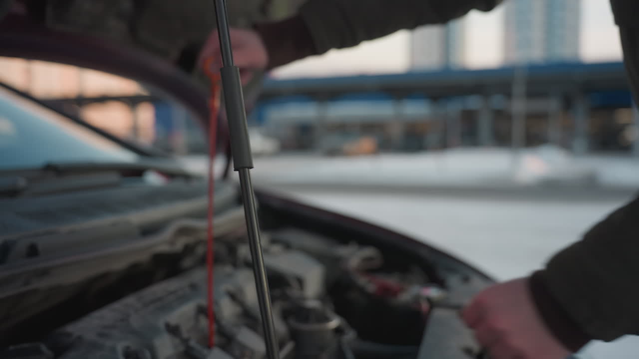 Close up of auto engineer in hooded jacket opening car hood to inspect oil gauge, checking oil level under cold winter conditions and closing it back, with urban background