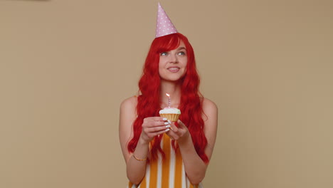 happy redhead girl celebrating birthday anniversary party, blowing candle on small cake cupcake