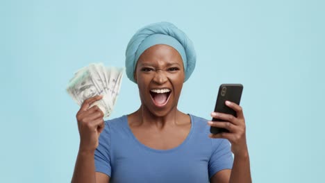 excited african american lady holding cellphone and money, blue background