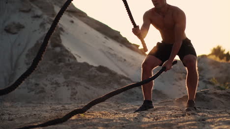 athletic man on exercise around the sand hills at sunset hits the rope on the ground and raised the dust.