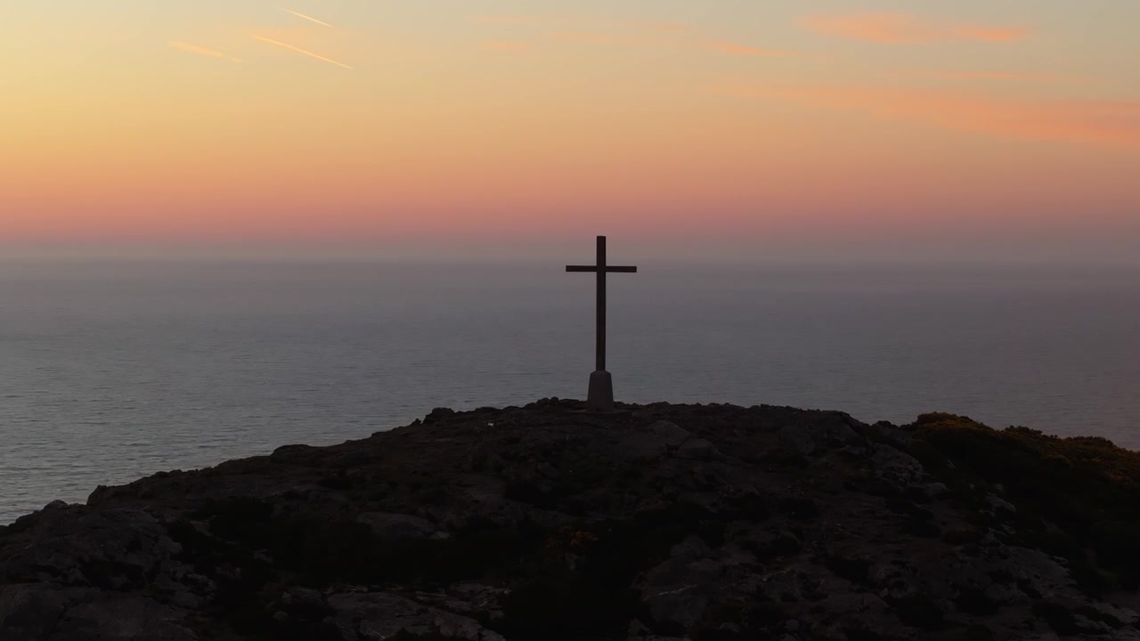 Una croce solitaria si erge su una collina, stagliandosi contro un cielo tenue all'alba con il mare calmo che si estende oltre. Ideale per temi spirituali o caratteristiche paesaggistiche costiere.