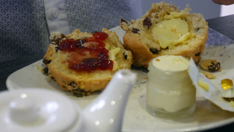 A close-up of a person spreading strawberry jam on a buttered scone, with clotted cream in a jar nearby