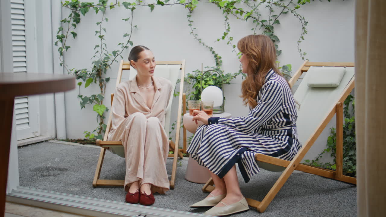 Two laughing women sitting apartment balcony at cozy chairs. Smiling girlfriends