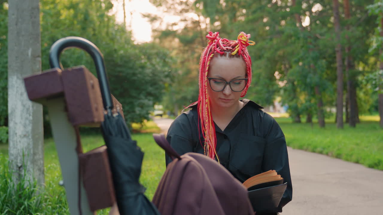 Mujer caucásica leyendo en un banco al atardecer, con trenzas rosas y gafas, novela abierta sobre su regazo, mochila colgada en el banco, luz dorada cálida proyectando largas sombras, sendero del parque bordeado de árboles, relajada y concentrada
