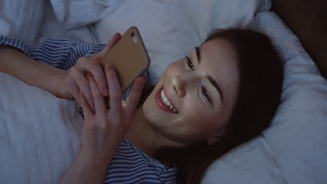 vista de cerca de la encantadora mujer caucásica con cabello oscuro acostado en la cama, charlando en el teléfono inteligente a altas horas de la noche